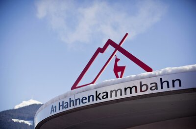 Ein Schild der Hahnenkammbahn mit einem stilisierten Dach und einem Reh. Der Schreibstil ist modern und ansprechend, vor einer bergigen Kulisse.