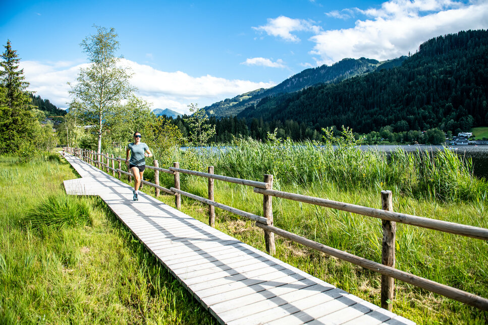Eine Läuferin joggt auf einem Holzsteg, umgeben von üppigem Grün und Bergen unter einem strahlend blauen Himmel.