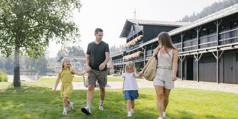Eine Familie mit zwei kleinen Mädchen spaziert fröhlich über eine grüne Wiese am Schwarzsee in Kitzbühel, umgeben von Bäumen und einem Gebäude im Hintergrund.
