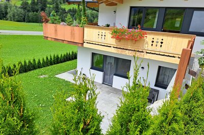 A modern house with a balcony, adorned with blooming plants. The garden is green and well-maintained, surrounded by a hedge.