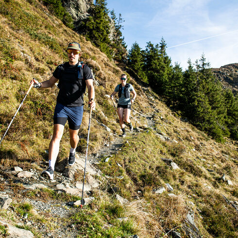 Zwei Trailrunner wandern auf einem schmalen Pfad durch eine malerische Berglandschaft mit grünen Bäumen im Hintergrund.