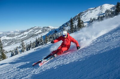 Skifahrer in rotem Anzug fährt eine präparierte Piste hinunter, im Hintergrund verschneite Berge und Nadelbäume.