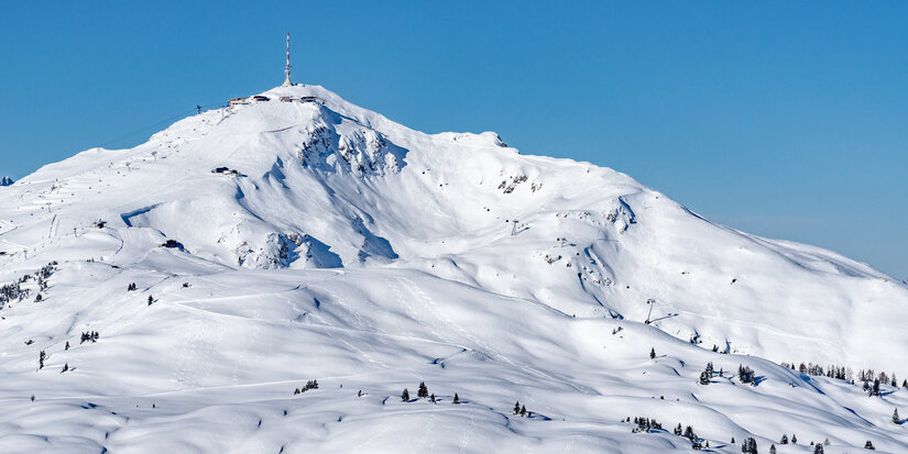 Am Kitzbüheler Horn im Skigebiet Kitzbühel erstreckt sich eine verschneite Berglandschaft unter strahlend blauem Himmel mit Aussichtsturm auf dem Gipfel.