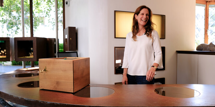 Eine lächelnde Frau steht an einem eleganten Tisch mit einer Holzbox, umgeben von kunstvollen Schmuckstücken in einem hellen Raum.