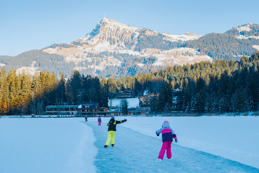 Zwei Kinder gleiten fröhlich über eine gefrorene Eisfläche, umgeben von schneebedeckten Bergen und einem klaren blauen Himmel.
