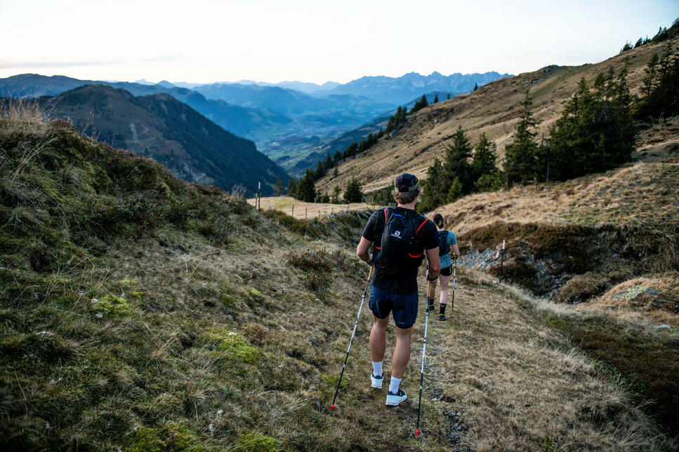 Drei Trailrunner wandern auf einem schmalen Pfad durch eine hügelige Landschaft mit Bergen im Hintergrund.