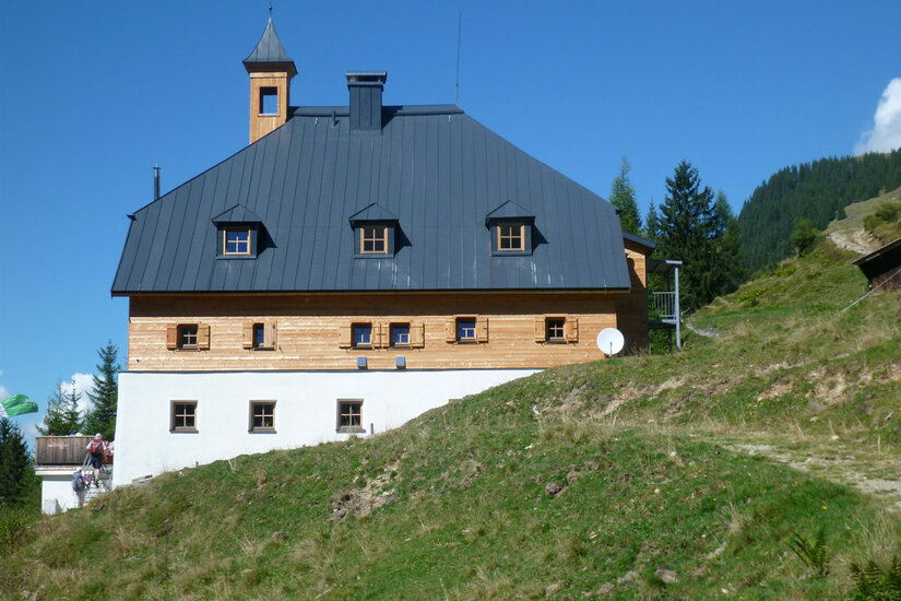 Dreistöckiges alpines Gebäude mit Holzfassade, weißem Erdgeschoss und dunklem Satteldach vor blauem Himmel.
