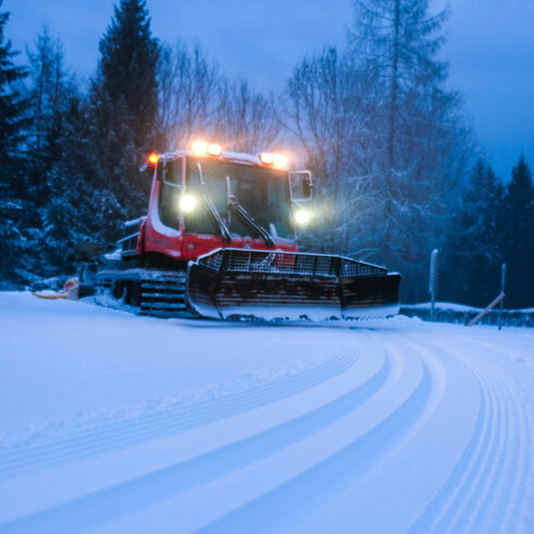 In Kitzbühel präpariert ein roter Pistenbully die Loipe in verschneiter Landschaft mit Tannen im Dämmerlicht.