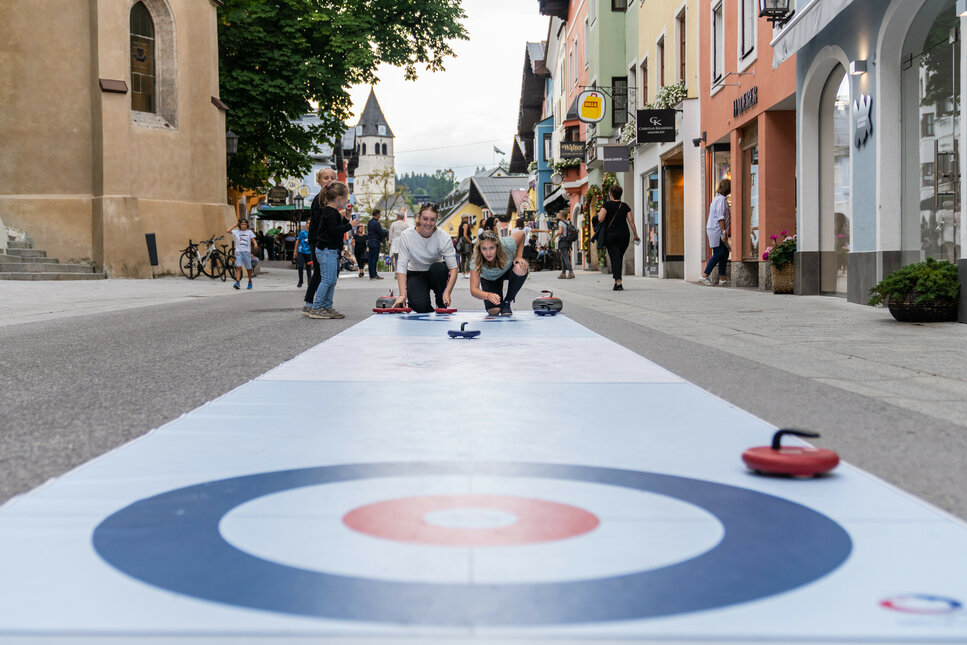Zwei Kinder spielen während Pura Vida in Kitzbühel auf einer bunten Curling-Bahn in einer belebten Straße, umgeben von Geschäften und Passanten.