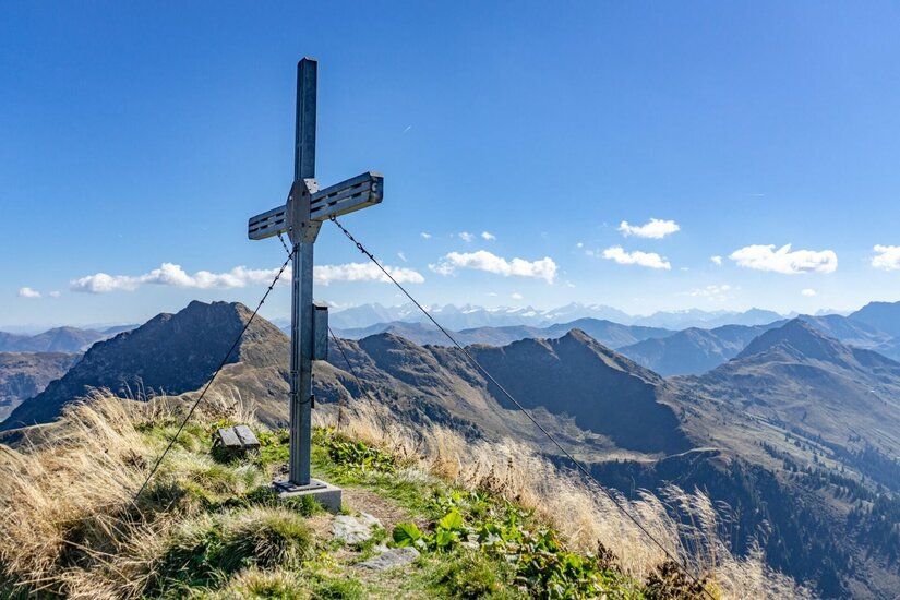 Gipfelkreuz an der Gebra Ein metallisches Gipfelkreuz auf einem grasbewachsenen Berggipfel vor einem Panorama aus weiteren Gebirgsketten.