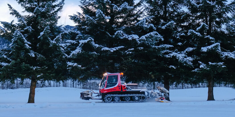 Beim Langlauf in Kitzbühel präpariert ein roter Pistenbully die Loipe in verschneiter Landschaft mit Tannen.