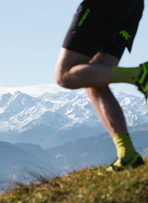 Der Fokus liegt auf dem Bein eines Sportlers, der in den Alpen joggt, während majestätische Berge die Landschaft prägen.