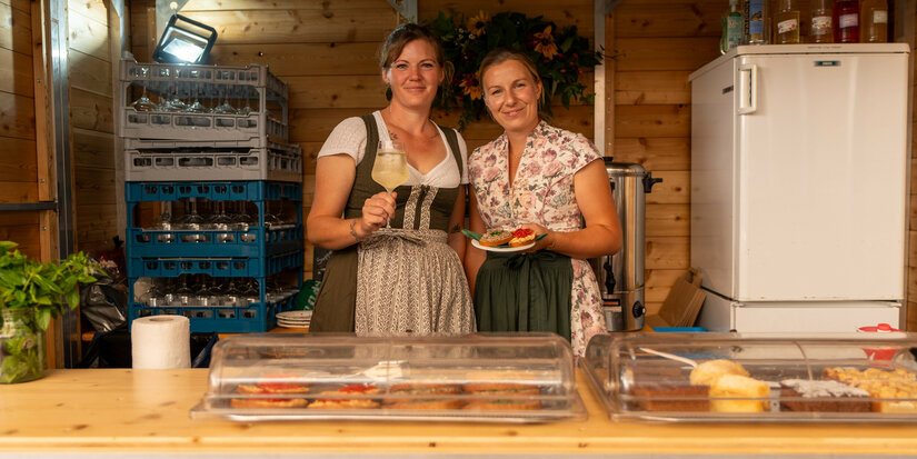 Zwei Frauen in Tracht stehen in einer Holzbaracke beim Kulinarik-Festival in Kitzbühel und präsentieren Getränke und Essen.