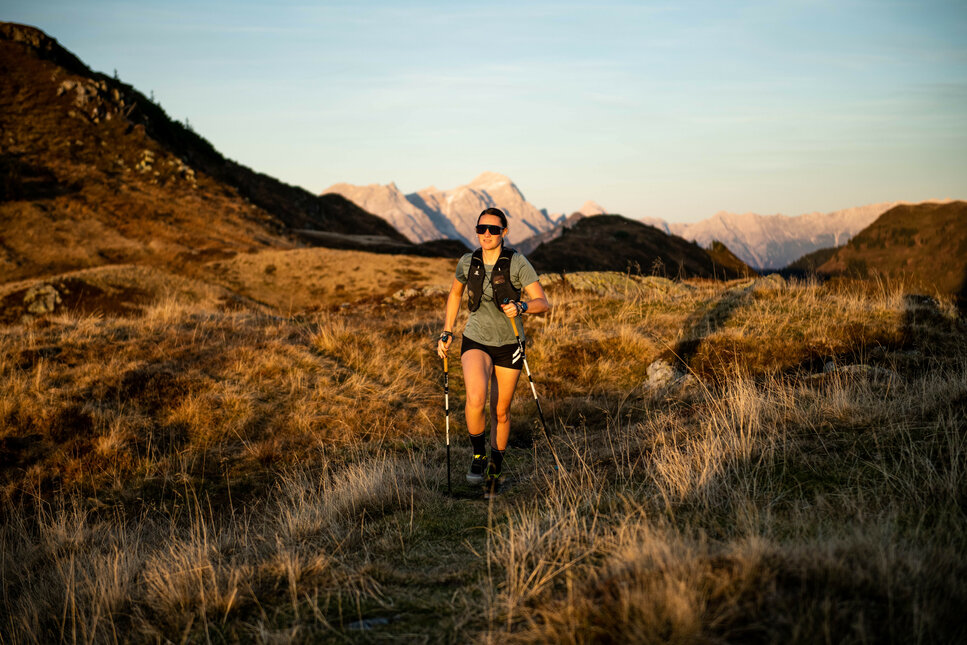 In der goldenen Abendsonne joggt eine Frau mit Sonnenbrille und Wanderstöcken durch die malerische Berglandschaft.