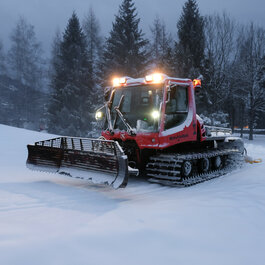 In Kitzbühel präpariert ein roter Pistenbully die Langlaufloipe in verschneiter Landschaft mit Tannen.