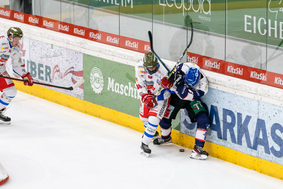 Zwei Eishockeyspieler kämpfen in Kitzbühel an der Bande um den Puck, während ein dritter Spieler im Hintergrund zuschaut.