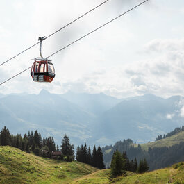 Eine rote Seilbahn der Hahnenkammbahn schwebt über die grünen Hänge des Hahnenkamms in Kitzbühel, umgeben von Bergen und einem bewölkten Himmel.