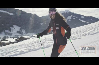 Skifahrerin in dunkler Skikleidung auf einer verschneiten Piste vor Bergpanorama.