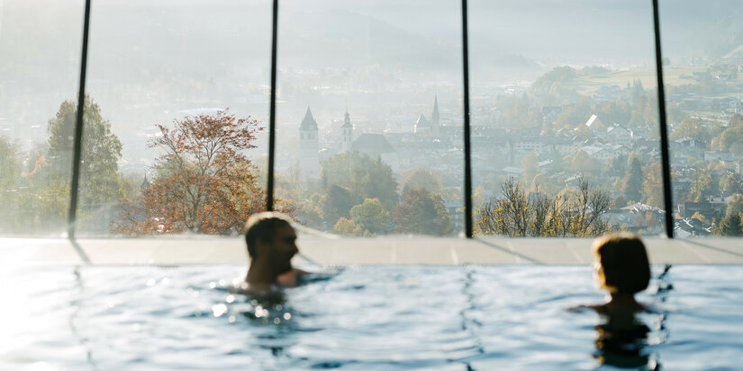 Im Vordergrund schwimmen zwei Menschen in einem Pool, während die malerische Stadt Kitzbühel im Hintergrund sichtbar ist.