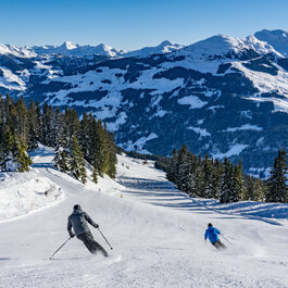 Jochberg, Skigebiet Kitzbühel, zwei Skifahrer auf verschneiter Piste mit Bergkulisse und Tannen.