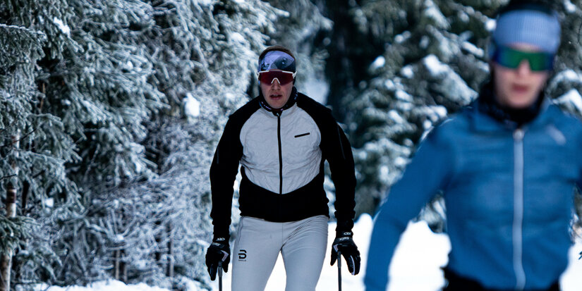Beim Langlauf in Kitzbühel gleiten zwei Langläufer durch verschneiten Wald mit hohen Tannen.