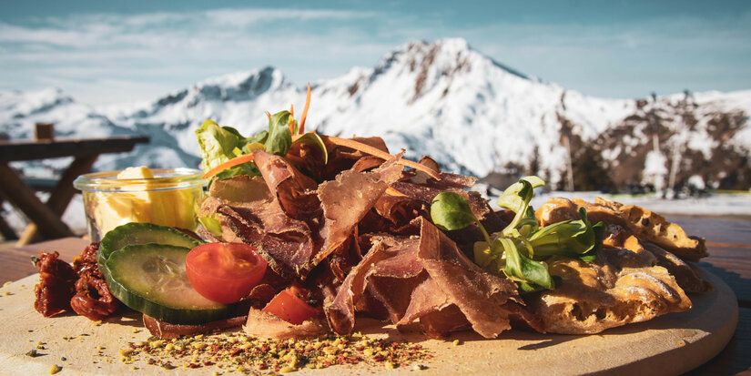 Bärenbadalm, Skigebiet Kitzbühel – Rustikales Holzbrett mit Schinken, frischem Gemüse und cremiger Sauce vor verschneiter Bergkulisse.