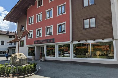 An outlet store in a two-story building with a red and brown facade. In front of the entrance is a large stone and a well-maintained green space.