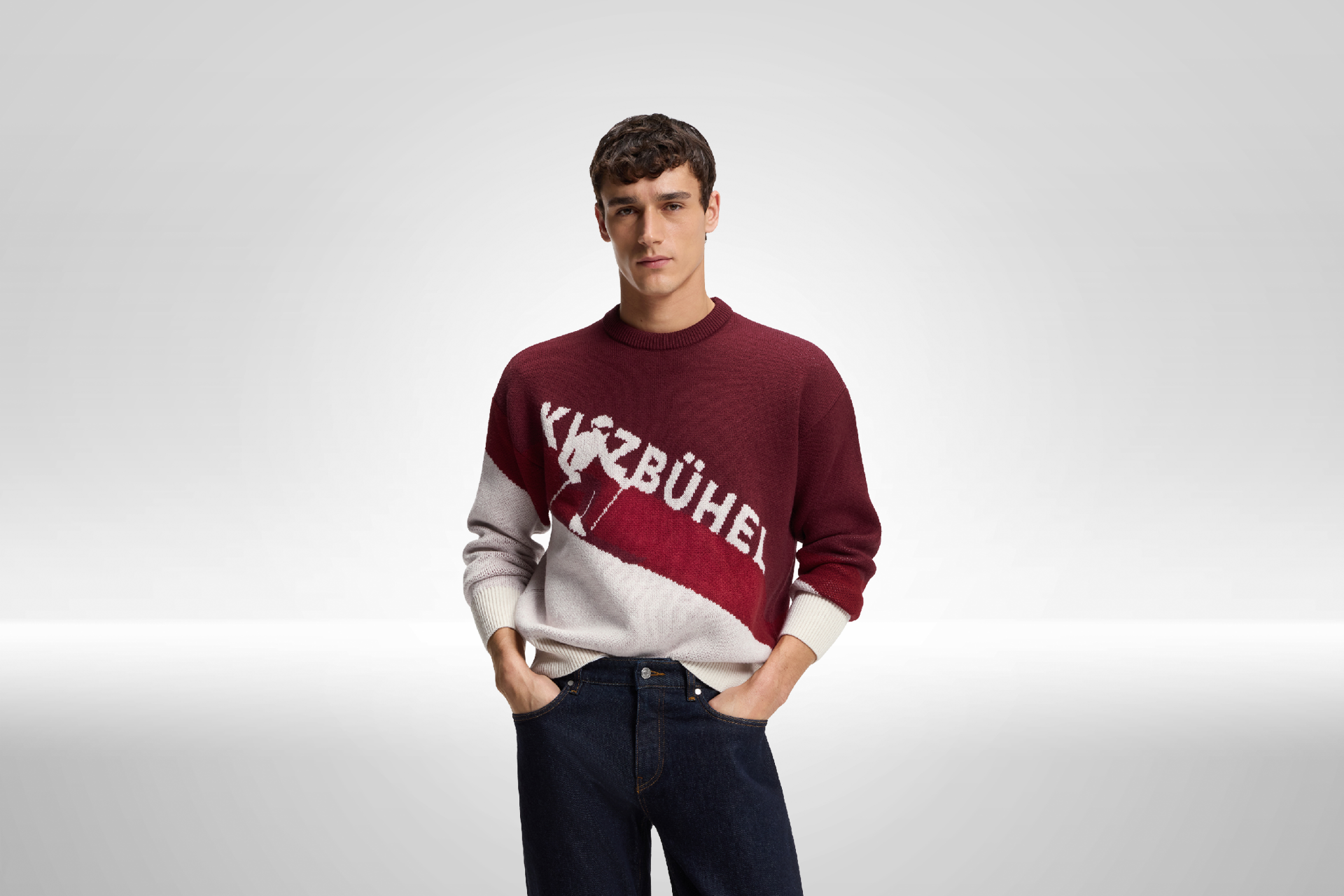 Ein junger Mann trägt einen rot-weißen Pullover mit dem Aufdruck 'Kitzbühel' und sieht selbstbewusst in die Kamera.