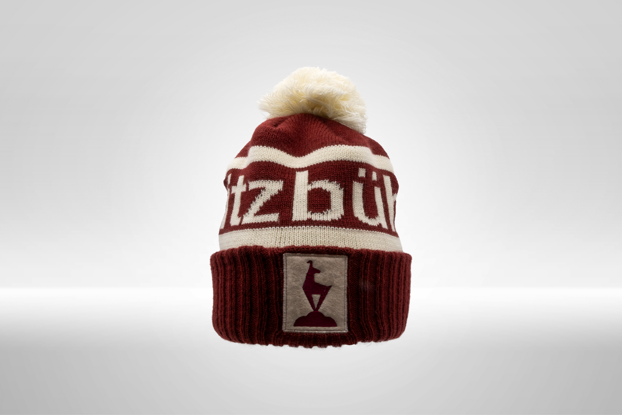 Eine dunkelrote Strickmütze mit einem weißen Schriftzug und einem flauschigen Bommel auf der Oberseite.